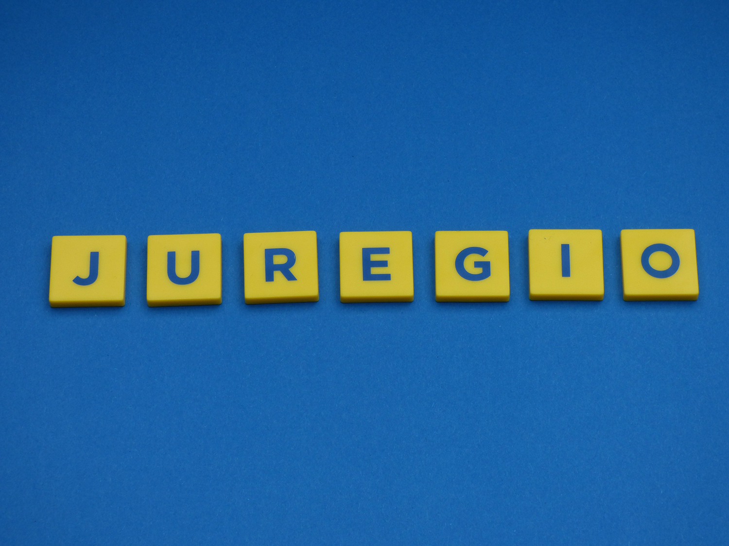 Auf einem blauen Untergrund ist das Wort JUREGIO aus sieben Buchstaben, welche sich auf einem gelben Hintergrund befinden, zusammengefügt.