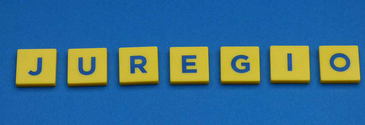Bild: auf einem blauen Untergrund ist das Wort JUREGIO aus sieben Buchstaben, welche sich auf einem gelben Hintergrund befinden, zusammengefügt.