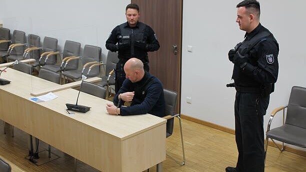 Bild: Justizwachtmeister bei der Bewachung während einer Gerichtsverhandlung. Sie stehen konzentriert hinter einem sitzenden Angeklagten.