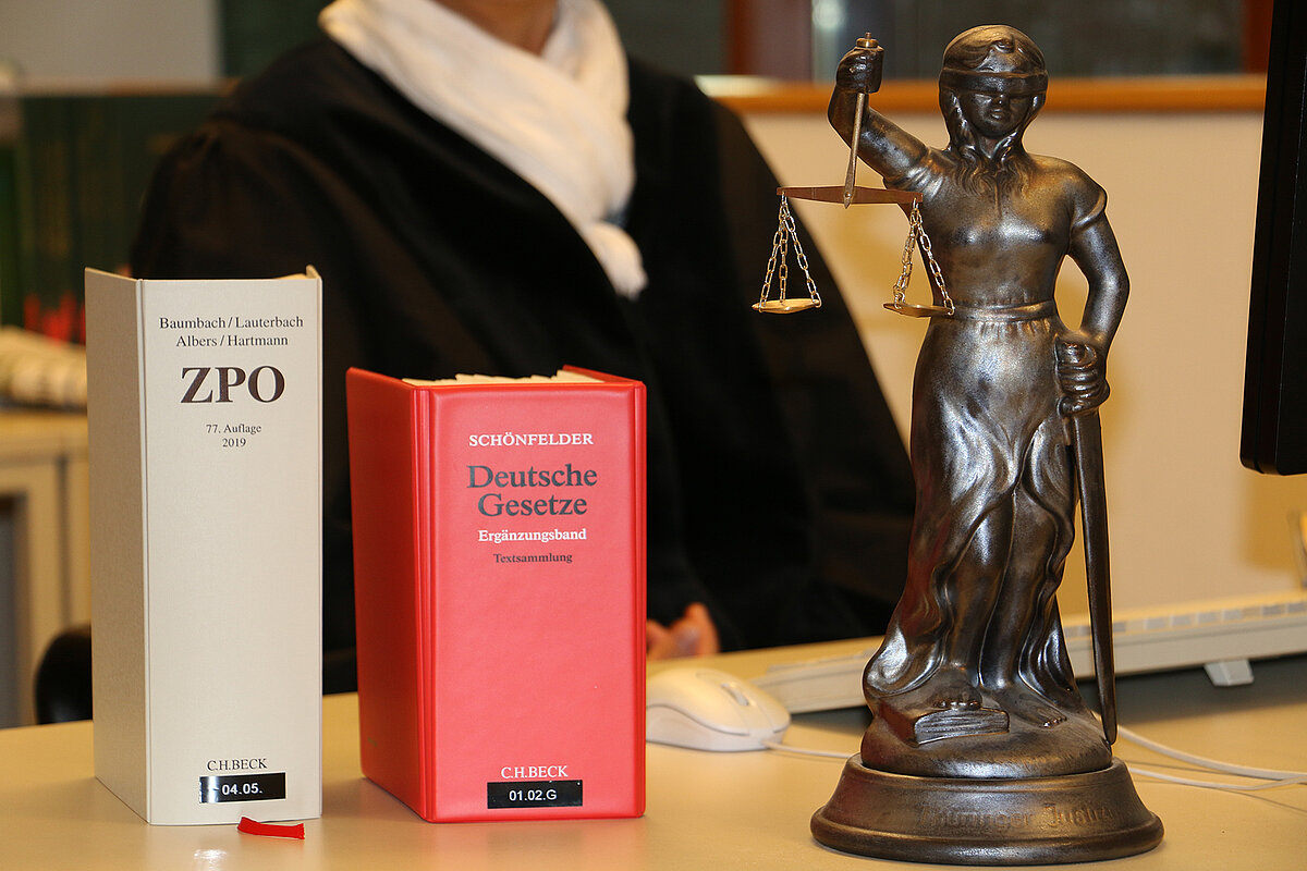 Bild: Gesetzestexte mit nebenstehender Justitia auf einem Schreibtisch